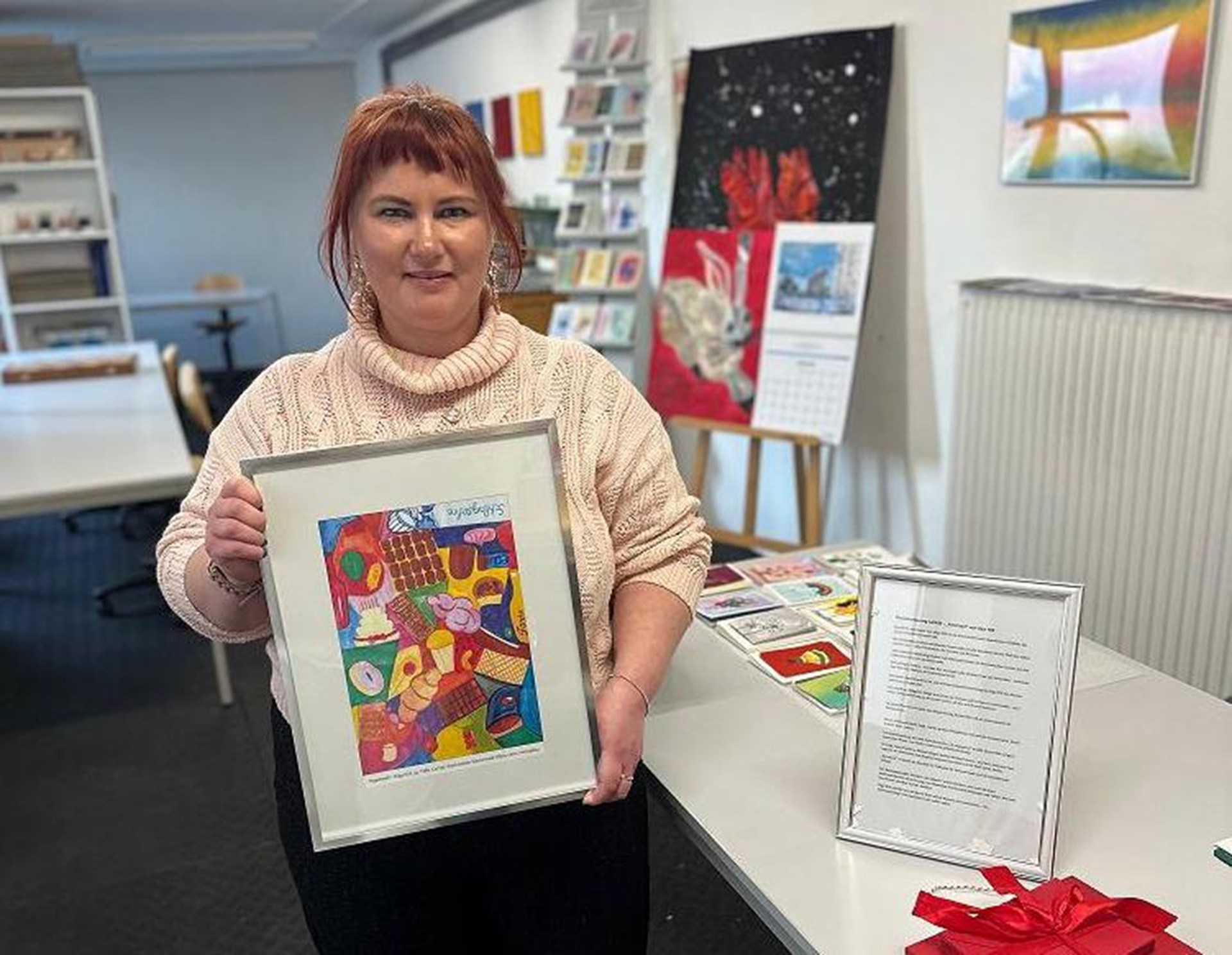 Eine Frau steht in einem Raum der Caritas-Werkstätten und hält ein gerahmtes, farbenfrohes Kunstwerk in den Händen. Auf einem Tisch neben ihr liegen Unterlagen und ein Geschenk. Im Hintergrund sind weitere Kunstwerke zu sehen.