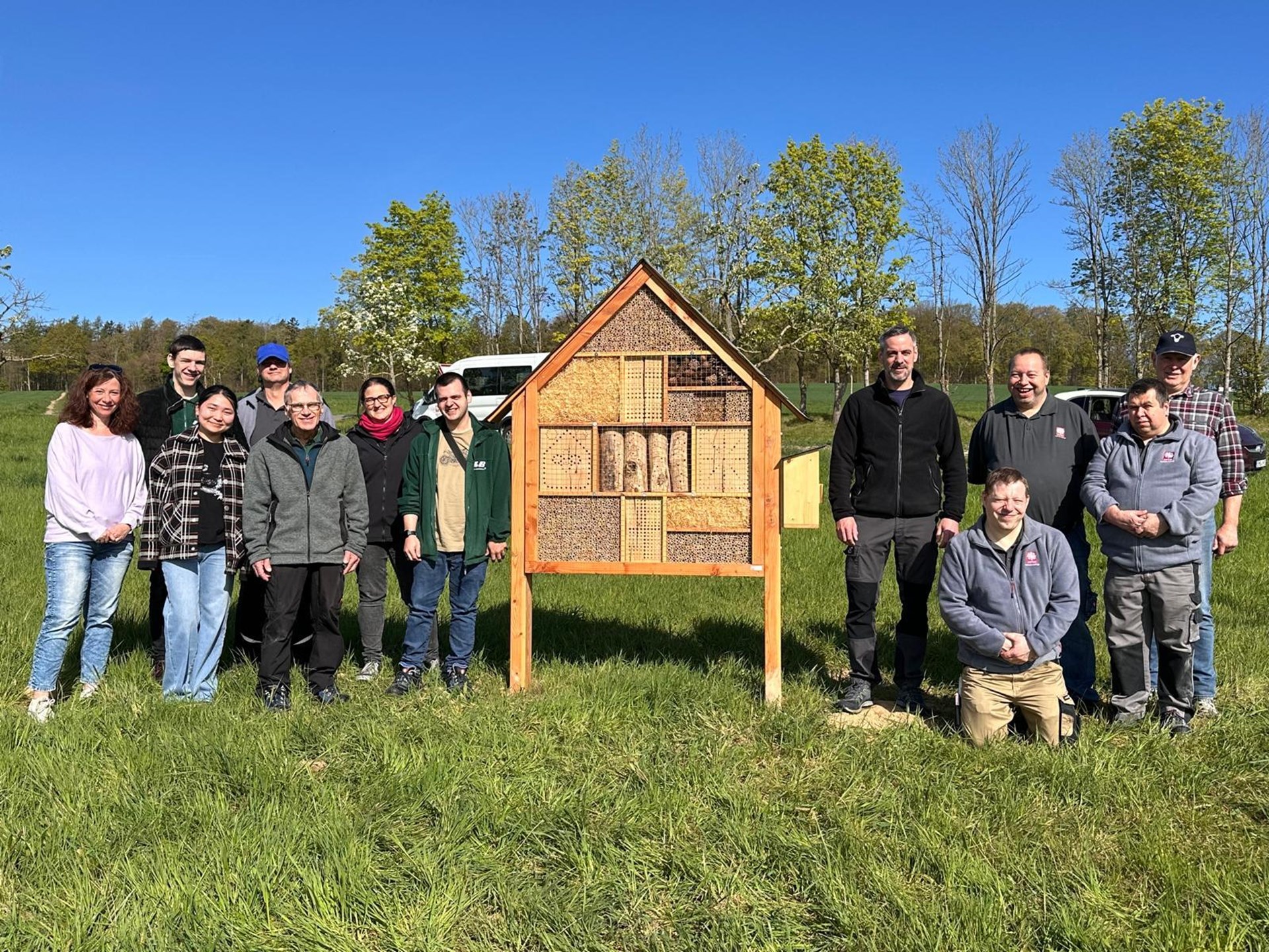 Gruppe von elf Menschen steht auf einer grünen Wiese rund um ein großes hölzernes Insektenhotel mit verschiedenen Naturmaterialien wie Holz, Bambus und Tannenzapfen. Im Hintergrund stehen Bäume unter einem klaren blauen Himmel an einem sonnigen Tag.