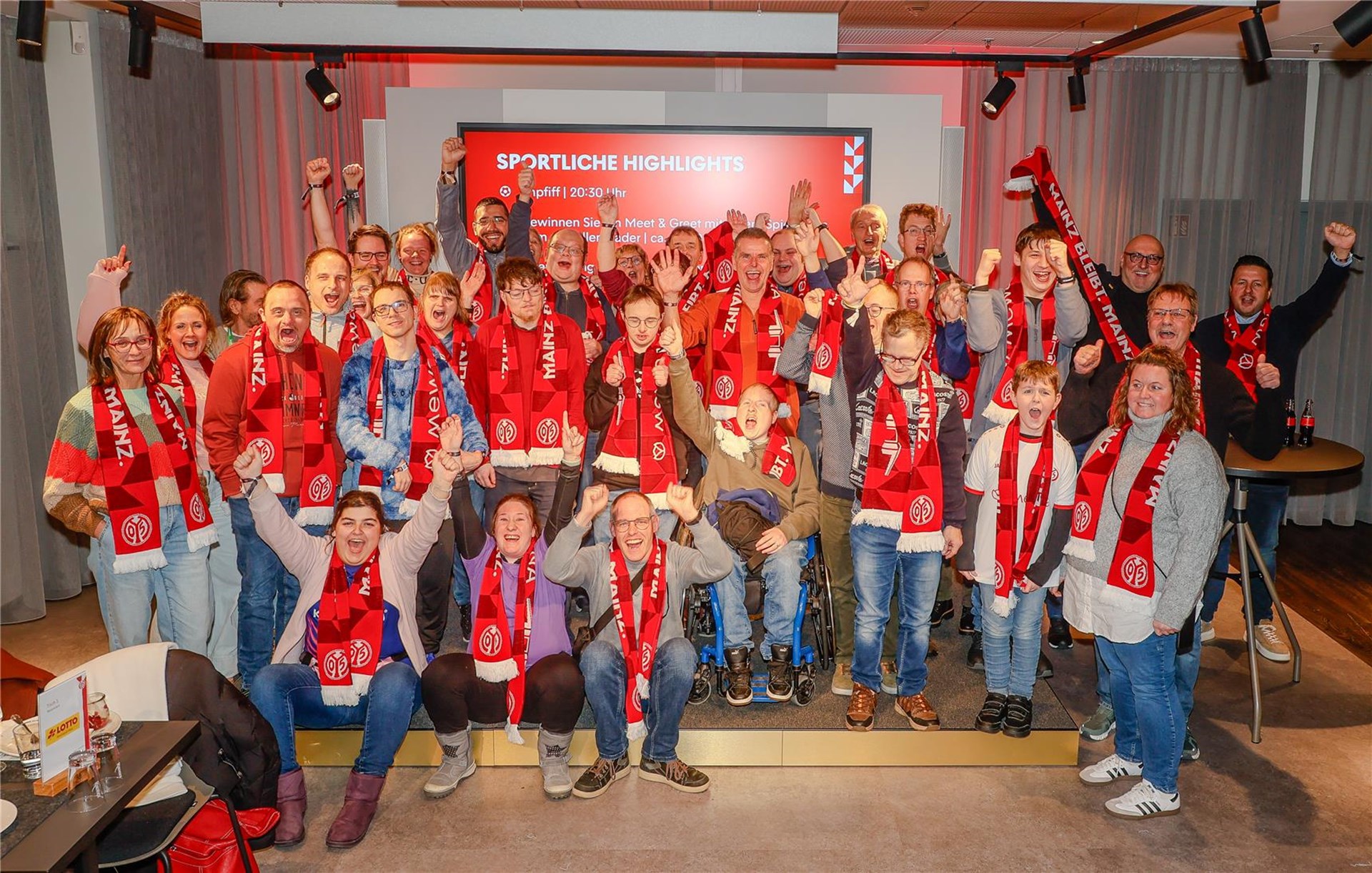 Eine große Gruppe von Beschäftigten und Mitarbeitenden der Caritas-Werkstätten Westerwald-Rhein-Lahn steht gut gelaunt im Stadion der MEWA Arena. Viele tragen rote Schals und lächeln in die Kamera. Im Hintergrund sind die Tribünen und das Spielfeld zu sehen.