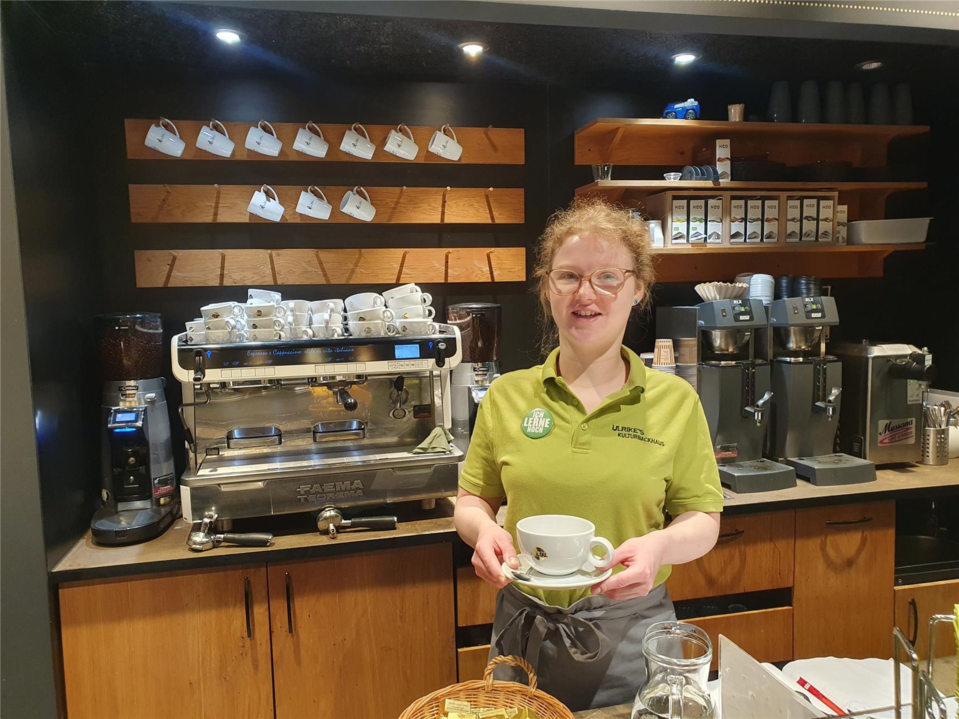 Eine Frau mit grünem Poloshirt und Brille steht lächelnd hinter der Theke eines Cafés. Sie hält eine Tasse, im Hintergrund eine Espressomaschine und Regale mit Tassen.