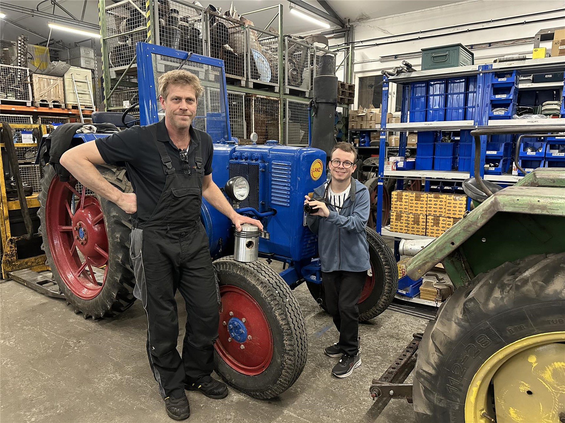 Ein Mann in Latzhose und ein jüngerer Mann stehen in einer Werkstatt neben einem blauen Lanz-Traktor. Sie lächeln, umgeben von Regalen mit Werkzeug und Ersatzteilen.